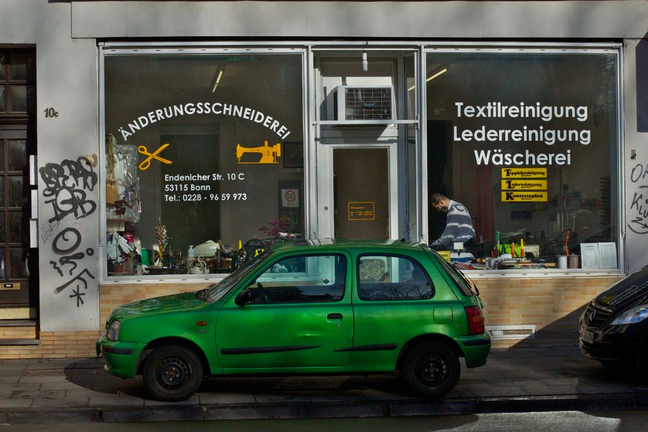 A storefront with large glass windows displaying text in German advertising fabric, leather, and laundry cleaning services. The shop has a brick base and a grey facade with multiple signs, including a yellow one for fabric cleaning and reparation. Inside, a person is visible working at a table with various cleaning supplies and tools. A small, bright green compact car is parked directly in front of the shop on the street, casting a shadow on the pavement. The scene is lit with natural daylight, highlighting the cleanliness of the surfaces and the organized interior, reflecting a professional approach to cleaning and maintenance. The image associates with residential and commercial cleaning, emphasizing surface cleaning, deep cleaning, and sanitisation services that Kennington Cleaners offers.
