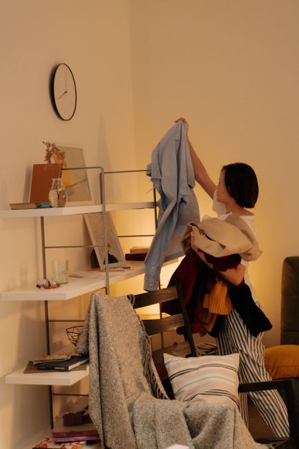 A woman in a white top and striped pants is folding laundry in a living room. She is holding a pile of clothes and reaching to hang a blue shirt on a metal clothes rack. The room features a white shelving unit with decorative items, a small clock on the beige wall, and a sofa with a cushion and a grey throw blanket. The surface of the coffee table below the shelves is clear, and the lighting is warm and soft, creating a cozy atmosphere. The scene depicts a domestic environment focused on routine cleaning and tidying activities, aligning with the context of general household maintenance and deep cleaning, such as surface cleaning and organisation, associated with Kennington Cleaners' services.