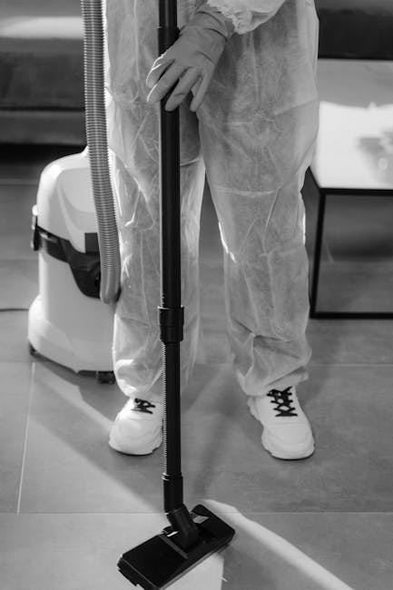 Close-up of a person in protective clothing, including gloves and a disposable suit, operating a vacuum cleaner with a wide cleaning head on a tiled floor in a commercial or domestic setting. The vacuum cleaner is connected to a hose and appears to be a commercial-grade device used for deep cleaning surfaces. The scene emphasizes surface cleaning and sanitisation processes carried out by Kennington Cleaners, highlighting their professional approach to maintaining hygiene and cleanliness in various environments. Bright, even lighting enhances the clarity of the surfaces and cleaning tools visible in the image.