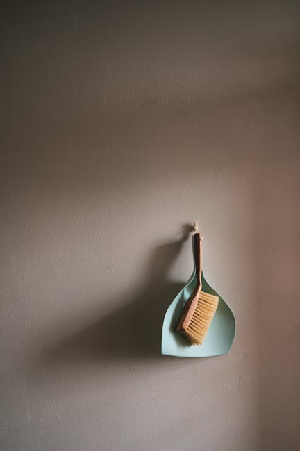 A close-up of a beige textured wall with a light blue plastic corner shelf mounted on it. Hanging from the hook on the shelf is a small, natural wood-handled cleaning brush with light-colored bristles. The surface appears clean and well-maintained, with no visible dust or dirt, illustrating an orderly domestic setting suitable for surface cleaning or polishing activities. The image emphasizes simplicity and cleanliness, highlighting tools used in standard household or commercial cleaning performed by Kennington Cleaners. The lighting is soft and natural, casting gentle shadows that accentuate the minimalistic arrangement. This visual aligns with the theme of domestic cleaning and hygiene maintenance discussed in the context of avoiding hidden charges in cleaning quotes on the [PAGE_TITLE] page of the kenningtoncleaners.org.uk website.