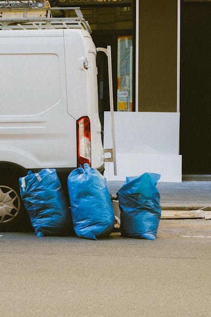 A white commercial van parked on a street next to a building with a large glass window and a white wall. The van's rear door is slightly open, revealing a clean, organized interior. Outside, three large blue plastic bags filled with cleaning supplies or waste are placed on the pavement near the back of the van. The pavement is smooth, and the scene is illuminated by natural daylight, highlighting the clean and well-maintained condition of the van and surroundings. This image exemplifies exterior commercial vehicle logistics related to surface cleaning and hygiene services, aligning with Kennington Cleaners' emphasis on professional cleaning and sanitisation, as referenced in the Kennington council permit guide for commercial cleaning vans.