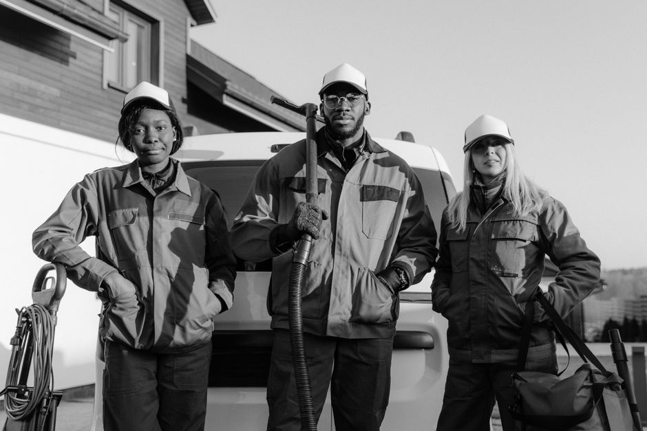 A group of three professional cleaning workers standing outdoors in front of a van, dressed in protective uniforms including jackets and caps. The central figure, a male with glasses and a beard, is holding a vacuum hose over his shoulder, while the woman on his left and the woman on his right are standing with one hand on their hips. All three are wearing gloves and appear prepared for surface cleaning and sanitation tasks. Visible cleaning equipment such as a vacuum cleaner and a bag are positioned next to them. The background features a modern building with large windows, and bright, natural lighting enhances the scene's clarity. This image exemplifies thorough domestic or commercial surface cleaning and sanitization practices, emphasizing hygiene and maintenance in line with [PAGE_TITLE], as provided by [COMPANY_NAME].