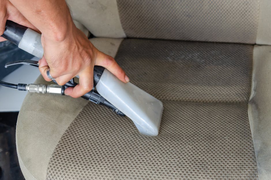 A close-up view of a person using a handheld steam cleaner on a beige fabric car seat cushion with a textured pattern, comparing the cleaned and uncleaned sections. The steam cleaner's nozzle is pressed onto the fabric, emitting steam to remove dirt and stains. The scene is well-lit, showing the contrast between the slightly stained and freshly cleaned areas, emphasizing surface cleaning and deep cleaning techniques. The background shows part of the car's interior, with the seat's backrest and side visible. The process highlights effective sanitisation and maintenance of upholstery, consistent with professional domestic or vehicle cleaning by Kennington Cleaners, a company specializing in carpet and upholstery cleaning.