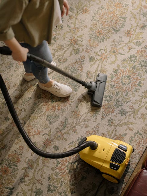 A close-up view of a person using a handheld steam cleaner on a beige fabric car seat cushion with a textured pattern, comparing the cleaned and uncleaned sections. The steam cleaner's nozzle is pressed onto the fabric, emitting steam to remove dirt and stains. The scene is well-lit, showing the contrast between the slightly stained and freshly cleaned areas, emphasizing surface cleaning and deep cleaning techniques. The background shows part of the car's interior, with the seat's backrest and side visible. The process highlights effective sanitisation and maintenance of upholstery, consistent with professional domestic or vehicle cleaning by Kennington Cleaners, a company specializing in carpet and upholstery cleaning.