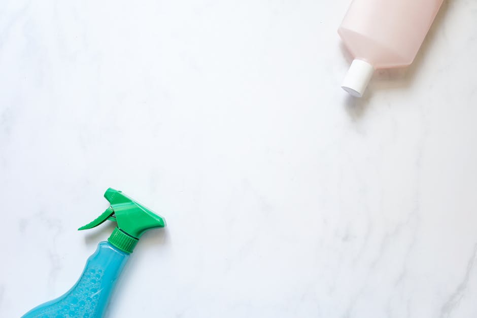 A light-colored marble or quartz countertop in a domestic kitchen with a clean, shiny surface. On the countertop, there is a green spray bottle and a blue cleaning cloth, suggesting recent or ongoing surface cleaning or sanitisation. In the background, a partially visible pink bottle, possibly containing cleaning solution, is placed toward the upper right corner. The lighting is bright and even, highlighting the reflective quality of the smooth surfaces. The scene emphasizes thorough cleaning processes typical of professional domestic cleaning services offered by Kennington Cleaners, aligned with their guide for tenants on surface cleaning and maintenance in residential environments.