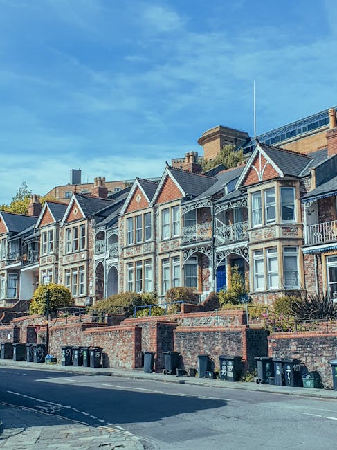 A row of Victorian-style terraced houses situated on a residential street, featuring brick facades with decorative white trim and ornate ironwork balconies. The houses have large bay windows, some with glass panels reflecting sunlight, and are set on brick foundations with landscaped front gardens. The street below includes a paved sidewalk, several black wheelie bins lined up along the curb, and is illuminated by natural daylight under a clear blue sky, creating a clean and well-maintained appearance. This scene represents the type of residential property that Kennington Cleaners specializes in for surface cleaning, deep cleaning, and sanitisation services in the Kennington area, as highlighted in the Kennington Road flat cleaning guide for tenants.