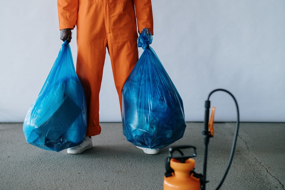 A person dressed in orange work overalls, visible from the waist down, is holding two large blue plastic garbage bags filled with waste. The bags are slightly translucent, revealing some of the contents inside. The individual is standing on a clean, grey concrete floor in a well-lit indoor space with a plain white wall background. A black and orange sprayer or pest control device is positioned on the floor in the foreground, slightly out of focus. The scene emphasizes waste disposal and sanitation practices, aligning with the requirements for surface cleaning and waste management policies, as recommended by Kennington Cleaners for maintaining hygiene compliance in residential and commercial settings.