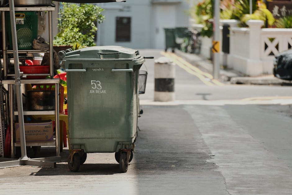 A green wheeled waste bin with the number 53 and the words 'JUN BESAR' is positioned on a paved sidewalk near a residential street in Lambeth, with a metal shelving unit holding cleaning supplies and tools to the left. The background shows part of a street with curb stones, bollards, and a black vehicle, with greenery and residential buildings further in the distance. The scene is outdoors during daylight, with natural lighting highlighting the clean, organized environment typical of surface cleaning and waste management areas, consistent with the principles discussed in Lambeth council rules for waste disposal and cleaning companies, as seen on the Kennington Cleaners website.