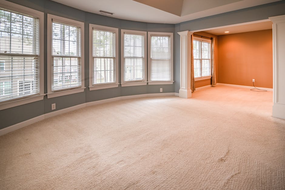 A spacious living room with a beige carpeted floor, featuring multiple large windows with white blinds allowing natural light to flood the space. The walls are painted in a soft gray, complemented by 