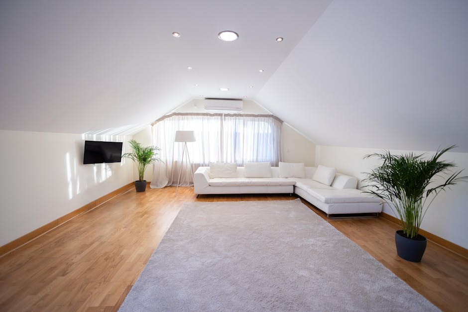 A spacious living room with a beige carpeted floor, featuring multiple large windows with white blinds allowing natural light to flood the space. The walls are painted in a soft gray, complemented by 
