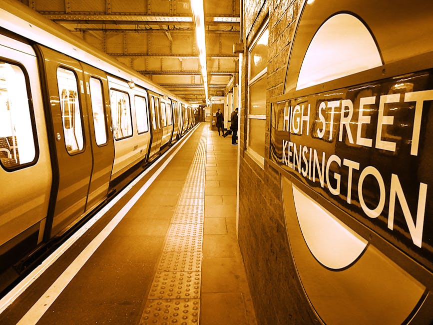 An underground railway station platform with a yellow and white train stopped alongside. The platform surface is tiled, appearing clean and dry, with tactile paving for accessibility running parallel to the train. The station's brick walls are illuminated by bright, overhead fluorescent lighting, creating a well-lit environment. On the right side, a roundel sign reads 'High Street Kensington,' indicating the station's location. A few passengers are visible in the background, waiting near the platform edge. The scene is a typical setting for surface cleaning and maintenance, emphasizing cleanliness and safety standards associated with domestic and commercial cleaning services offered by Kennington Cleaners. The overall appearance is tidy, with all surfaces clear of dust and debris, reflecting effective surface cleaning and hygiene practices in a public transportation environment.