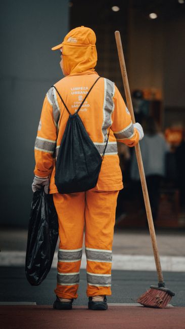 A professional cleaner from Kennington Cleaners wearing bright orange high-visibility workwear, including a jacket, pants, and a cap, stands outdoors on a paved surface holding a wooden-handled broom in their right hand and a black garbage bag in their left. The cleaner has a black bag slung over their shoulder and appears prepared for street or public space cleaning. The background is blurred but hints at a modern indoor environment with warm lighting, suggesting a focus on maintaining cleanliness and hygiene in commercial or residential areas through surface cleaning and waste removal. The scene emphasizes the importance of thorough sanitation and maintenance work.