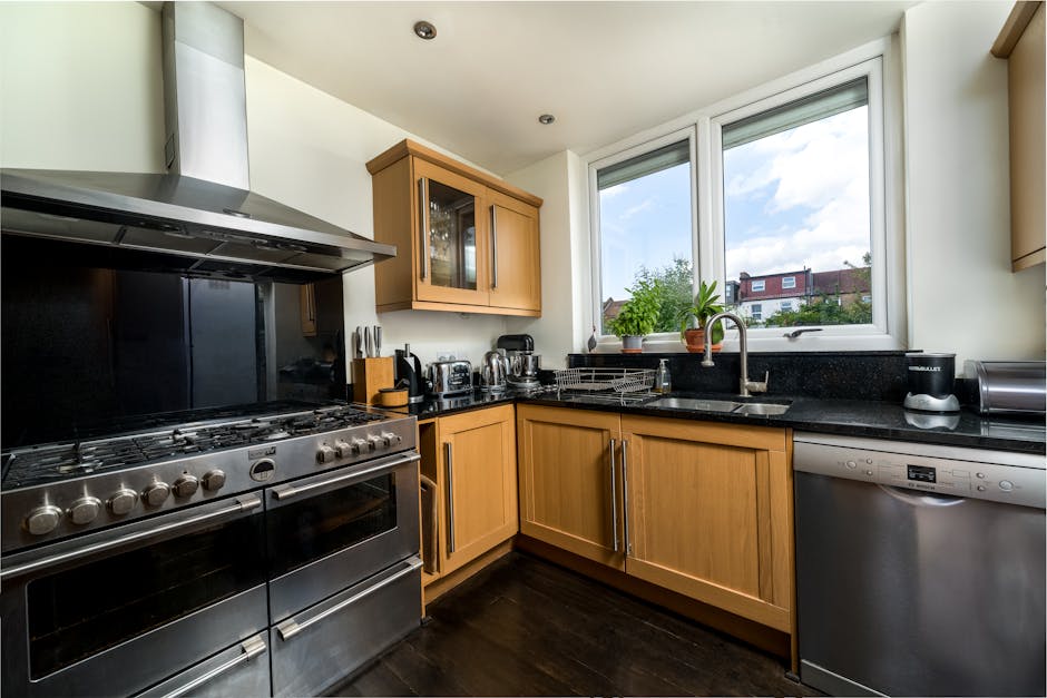 A modern kitchen scene featuring wooden cabinetry with a light finish, a stainless steel gas stove with multiple burners, and a black granite countertop. The countertop has a stainless steel sink with a silver faucet, and a small potted plant is placed near the window. The window allows natural light to illuminate the space, revealing a view of neighboring houses and blue sky outside. Visible kitchen appliances include a stainless steel toaster, a kettle, and a dishwasher. The surfaces appear clean and well-maintained, with no visible dust or dirt, highlighting the high standards of surface cleaning and sanitisation provided by Kennington Cleaners for residential properties in the SE11 area as part of their specialist cleaning services.