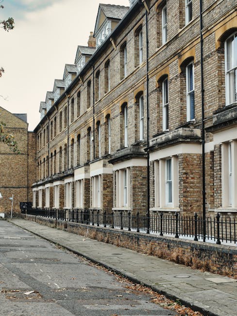 A row of traditional brick residential buildings with white-framed windows and black wrought iron railings along the sidewalk. The pavement appears clean, with some fallen leaves scattered on the ground. The exterior walls of the buildings show a weathered brick surface, while the windows are spotless and reflective, suggesting recent cleaning or good maintenance. The scene is illuminated by natural daylight, highlighting the texture of the brickwork and the neatness of the surroundings. This setting illustrates the type of property that Kennington Cleaners provides surface cleaning and deep cleaning services to, ensuring hygiene and pristine presentation for residential and commercial spaces in the SE11 area, in accordance with landlord cleaning requirements for Kennington properties.